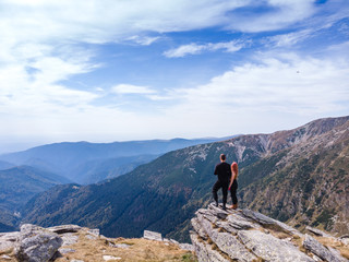 Fototapeta premium Back view of tourist couple, man and girl stand on rocky mountain top enjoying breathtaking autumn mountain panorama. Tourism, traveling and healthy lifestyle concept. motorcyclists