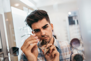 Handsome young man choosing eyeglasses frame in optical store.