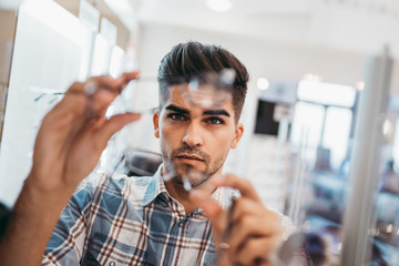 Handsome young man choosing eyeglasses frame in optical store.