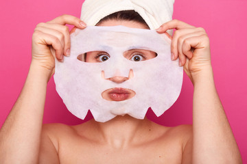 Close up of funny woman looks through white paper mask, has facial treatments, wears towel on head,...