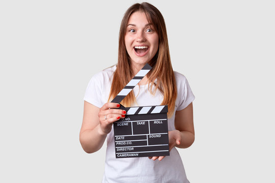 Film productivity concept. Satisfied female with long hair, looks with happy expression, dressed in white t shirt, holds clapper board, has red manicure, prepares for shooting new scene of movie