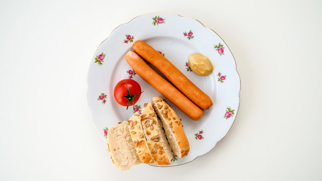 Par&oacute;wki, bułka, pomidor, musztarda na białym talerzyku leżące na białym tle