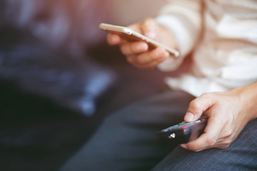 young man using television remote control and mobile smart phone  is sitting on a sofa. on or off tv. 