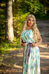 Young beautiful blonde woman in white dress with blue and green flowers posing in forest in summer