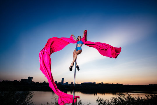 Young slim woman pole dancing at night on sky and city background