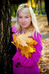 Young blonde woman in violet coat in park at autumn with yellow foliage