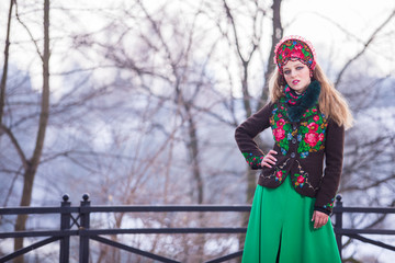 Fototapeta na wymiar Portrait of Beautiful Caucasian Girl in Fashionable Green Dress and Kokoshnik with Flowery Pattern and Beads. Posing in Winter Snowy Park