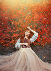 Fototapeta premium warm art photo of sleeping beauty, girl with fiery red hair lies on ground in dense forest under orange leaves in bright yellow sunlight, wonderful enchanted princess, new story about Snow White