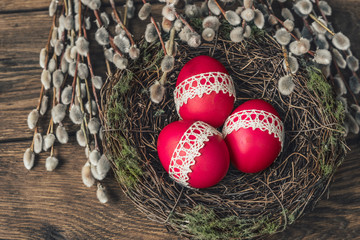 Red Easter eggs, decorative nest and willow branches against dark wooden background.