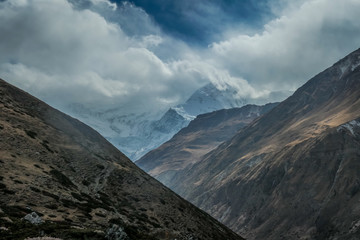 A barren mountain slopes covered with clouds. Mountains in the back covered with thick layer of snow. Annapurna Circuit Trek, Himalayas, Nepal. Harsh and steep slopes. Dangerous weather condition.