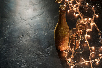 Photo of golden champagne bottle, two wine glasses, burning garland on black background