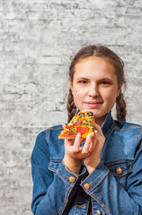 portrait of young teenager brunette girl with long hair eating slice of pizza on gray wall background with copy space.