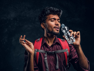 Handsome Indian hiker with backpack exhausted by heat gladly drinks refreshing water. Studio photo against a dark textured wall