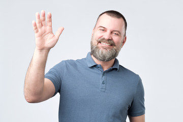 Polite ymature man with beard dressed in denim shirt saying hi, waving with hand