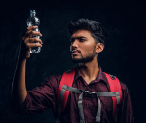 Handsome Indian hiker with backpack holding a bottle of water looking at her. Studio photo against a dark textured wall