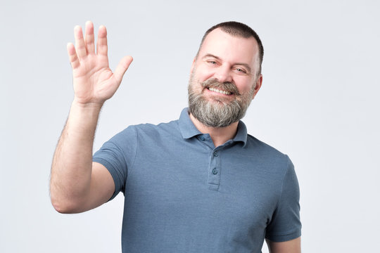 Polite ymature man with beard dressed in denim shirt saying hi, waving with hand