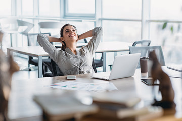 Happy businesswoman relaxing with hands behind head at office desk. Daydreaming concept