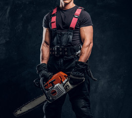 Handsome young lumberjack wearing protective clothes posing with a chainsaw in a dark studio
