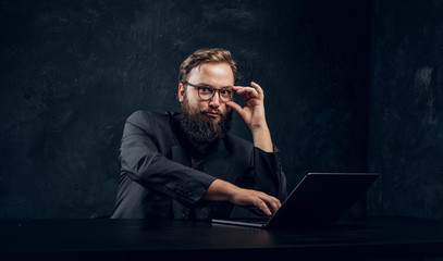 Bearded manager working behind a laptop and a cunning look at the camera holding glasses in the...