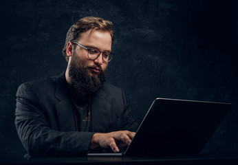 Bearded programmer in suit working on laptop sitting at the table in office against a dark wall