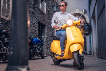 Fototapeta na wymiar Fashionable man wearing sunglasses riding on vintage Italian scooter in the old narrow street of Europe