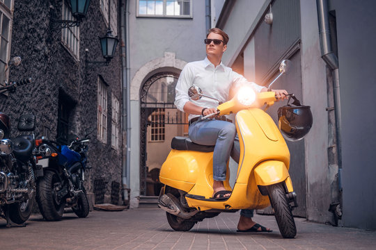 Fashionable man wearing sunglasses riding on vintage Italian scooter in the old narrow street of Europe