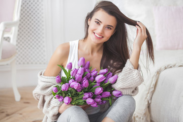 Attractive young woman with flowers indoors in the bedroom. Portrait of beautiful lady at home. Close up shot of female with tulips.