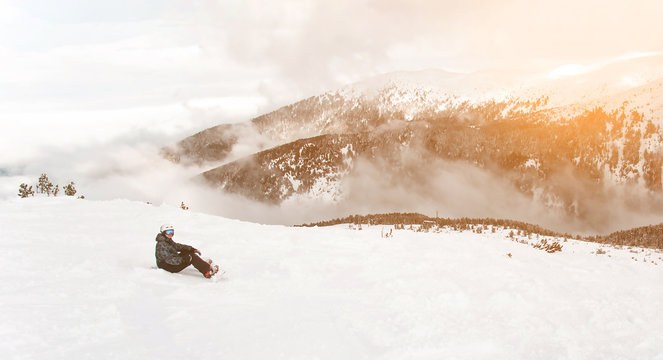 Panorama with snowboarder sit with snowboard in hands sits on big rock on mountains view. Bansko, Bulgaria
