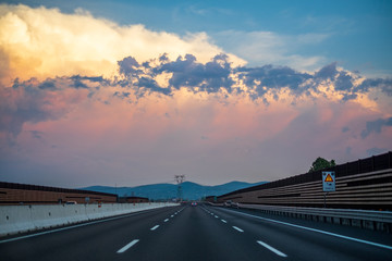 Sunset in clouds with sunrays over road to horizon. Travel background. Highway at mountains, beautiful landscape in Italy