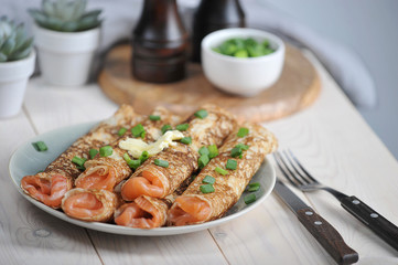 Pancakes with salmon. Pancakes sprinkled with green onions. Next to the plate are cutlery. Light wooden background. Close-up. 