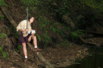 Girl in a Scottish kilt and animal skin on his shoulder posing in forest holding a Viking sword