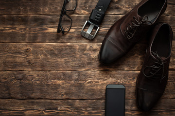 Businessman accessories such as mobile phone with a blank screen, glasses, belt and a elegant shoes on a brown wooden floor background with copy space.