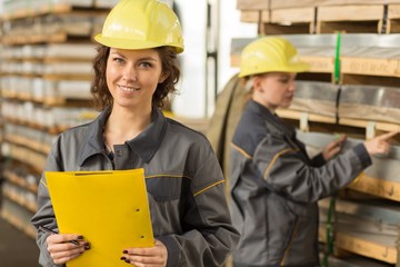 Two female engineers standing and checking material in storehouse. Women wearing yellow helmets and...