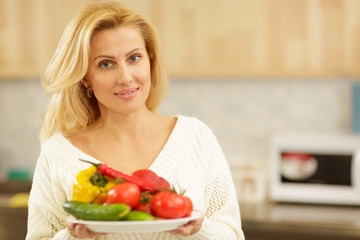 Obraz na płótnie Canvas Beautiful happy healthy mature woman smiling to the camera posing at her kitchen with a plate full of delicious vegetables. Housewife preparing healthy diet meals at home