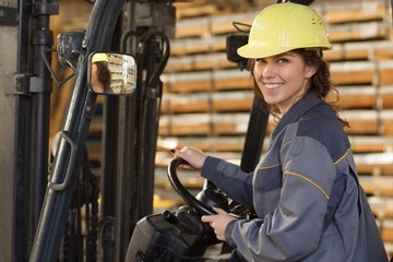 Woman industrial worker in gray uniform and yellow helmet driving by forklift truck, leaning...