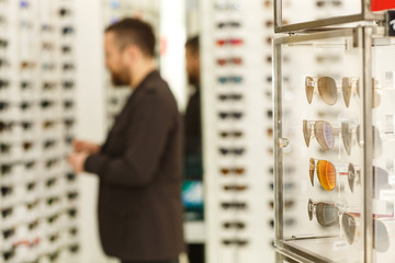 Selective focus on a showcase with sunglasses for sale on the foreground, copy space. Male customer choosing eyewear from the display, shopping at optical store