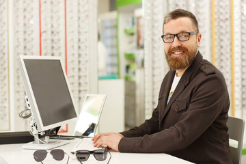 Horizontal portrait of a cheerful mature male professional optometrist working at his eyewear store. Handsome male optician smiling to the camera, using his computer