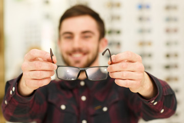 Cheerful handsome an holding out eyeglasses to the camera, standing at optics store. Happy male optometrist offering new glasses to the viewer. Eyesight, vision, eyes, health concept