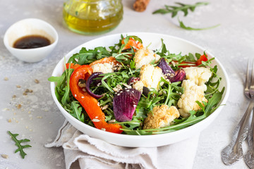 Salad with arugula, roasted vegetables (cauliflower, red bell pepper and onion) and seeds. Healthy food. Light grey concrete background. Copy space.