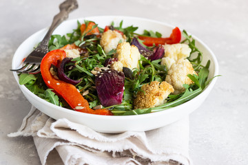 Salad with arugula, roasted vegetables (cauliflower, red bell pepper and onion) and seeds. Healthy food. Light grey concrete background. Copy space.