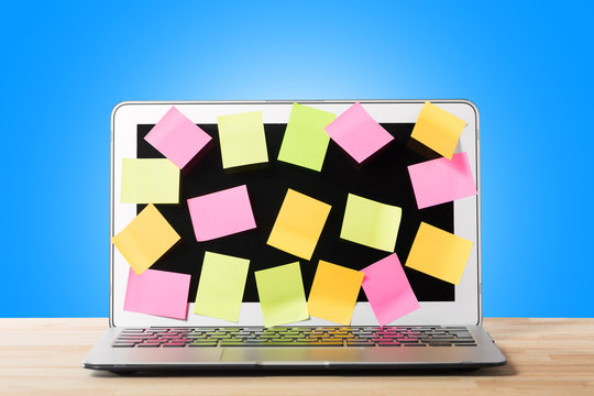 Many blank sticky notes covering modern laptop screen on wooden table against blue background. Concept of deadlines or ideas.