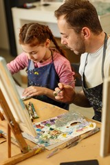 Family of brunet father with beard and red haired girl drawing picture together and mixing oil paints on palette. Father and pretty little daughter against oil picture on table. Private lessons of art