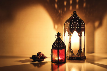 Ornamental Arabic lanterns with burning candles glowing at night. Plate with date fruit on the table. Festive greeting card, invitation for Muslim holy month Ramadan Kareem. Iftar dinner background. - obrazy, fototapety, plakaty