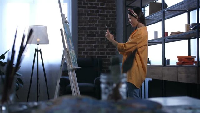 Side view of young asian woman artist taking a picture of her masterpiece at easel on cellphone in domestic art studio. Charming painter sharing her artwork with friends in social media