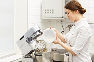 Pretty chef cooking cream for delicious  desserts and cakes using special modern kitchenware. Beautiful and handsome girl putting sugar in metal bowl of electric mixer.
