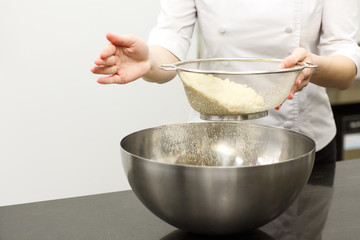 Hands of female chef holding sieve and sowing flour into big metal bowl for dough. Confectioner wearing white uniform. Concept of cooking desserts, confectionery and cuisine.