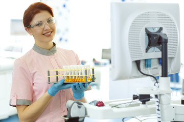 Smiling female lab technician with ginger hair standing in laboratory and posing. Lab worker wearing pink uniform, blue gloves and protective spectacles. Woman holding test tubes.