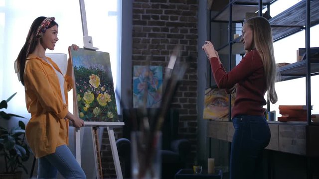 Joyful asian female artist posing to her friend with her masterpiece of yellow peonies. Caucasian smiling fan of painter's talent making photo of artist with new canvas on easel in home art studio