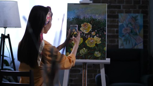 Back view of woman artist taking photo of finished masterpiece on smartphone in home art studio. Female painter photographing artwork of lage yellow peonies