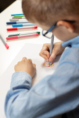 Chłopiec w niebieskiej koszuli i okularach siedzi przy biurku i rysuje na kartce papieru kolorowymi flamastrami.